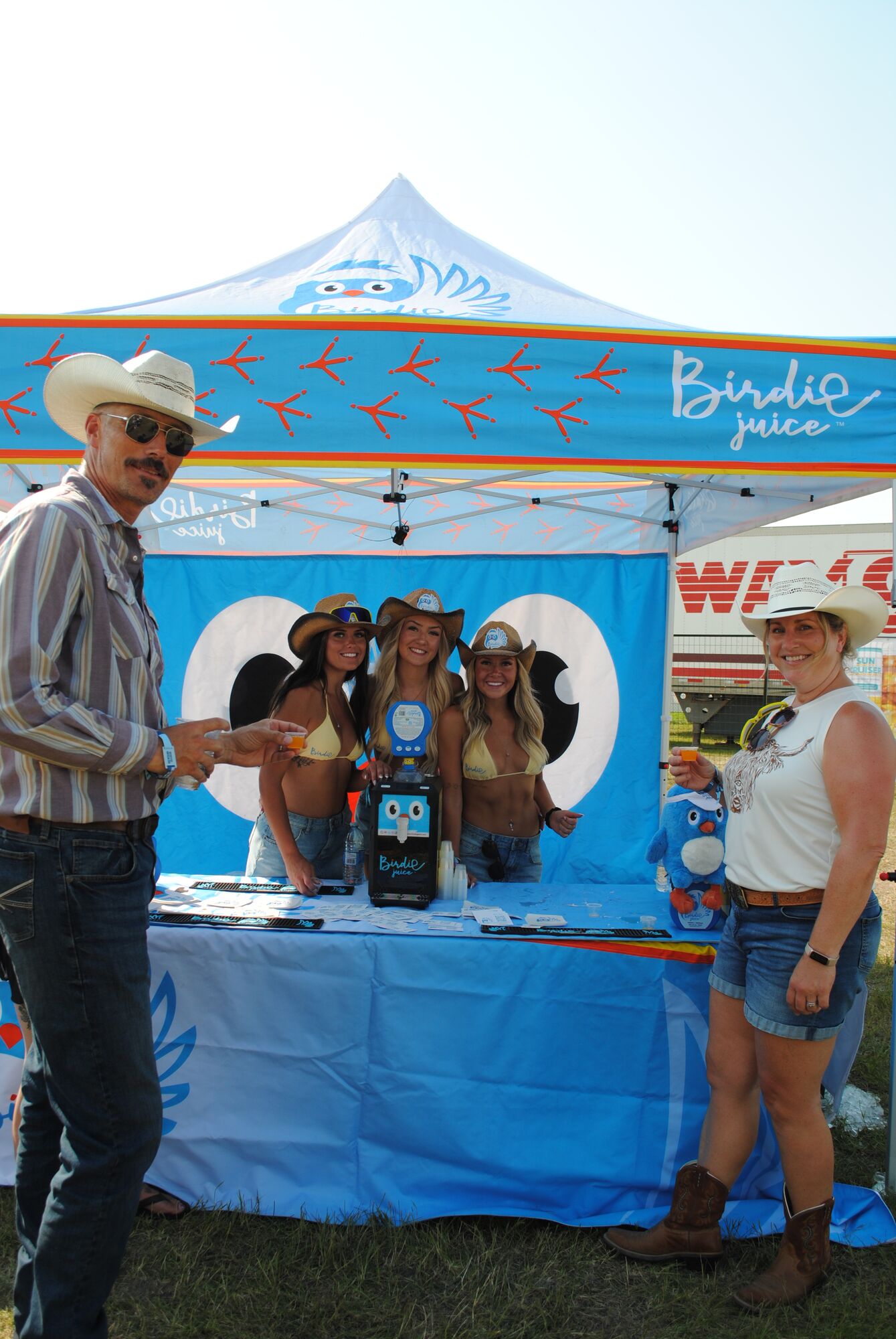 The Birdie Juice festival booth under a branded blue canopy, with the crew serving samples across a bottle-lined counter